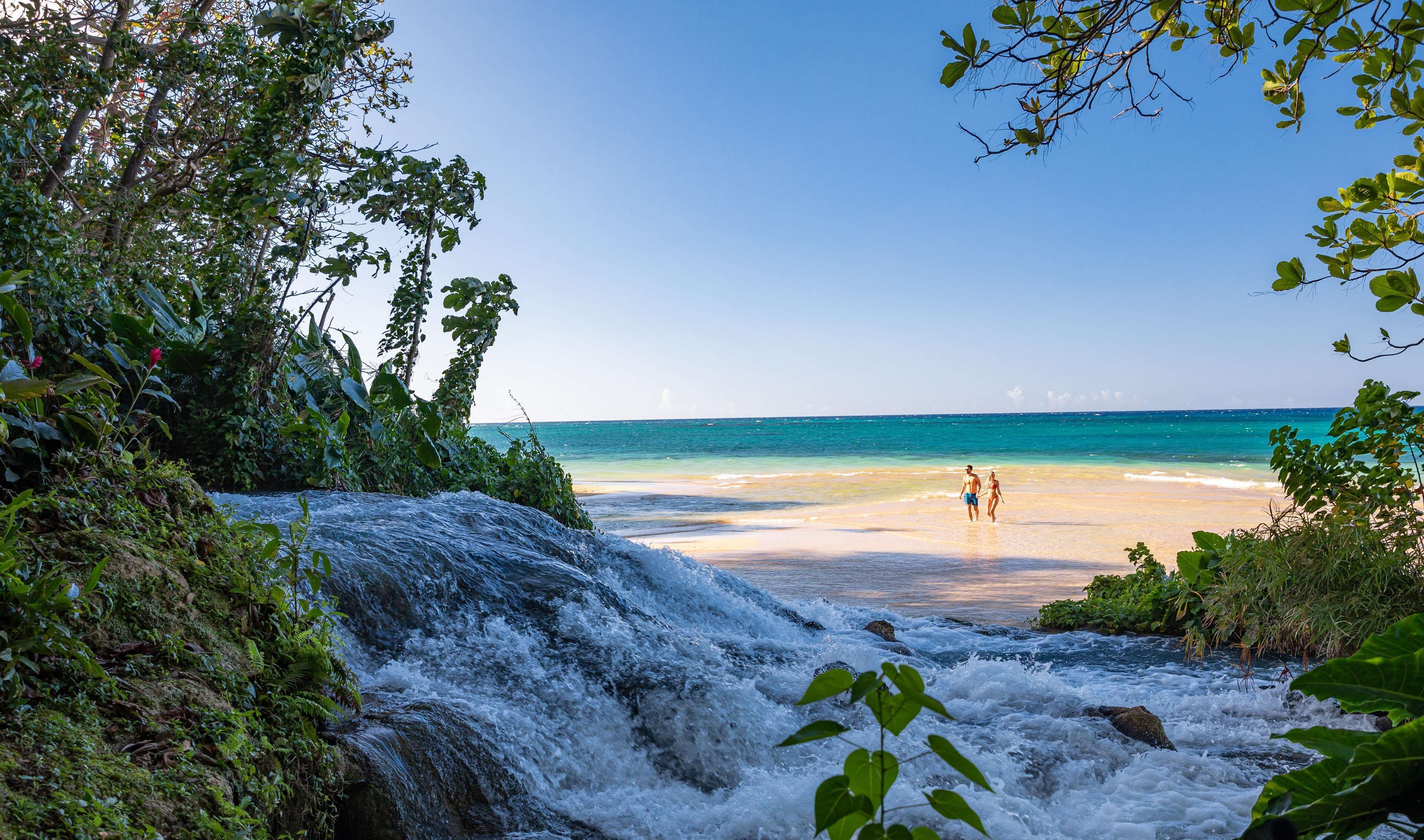 A waterfall leading to a beach with clear blue water and a couple walking on the shore.