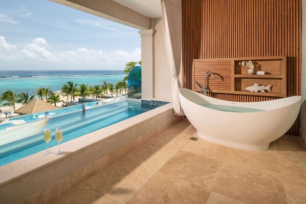 Un baño lujoso con una bañera grande, un lavamanos y un tocador de madera. Se ve una piscina y una playa con palmeras.