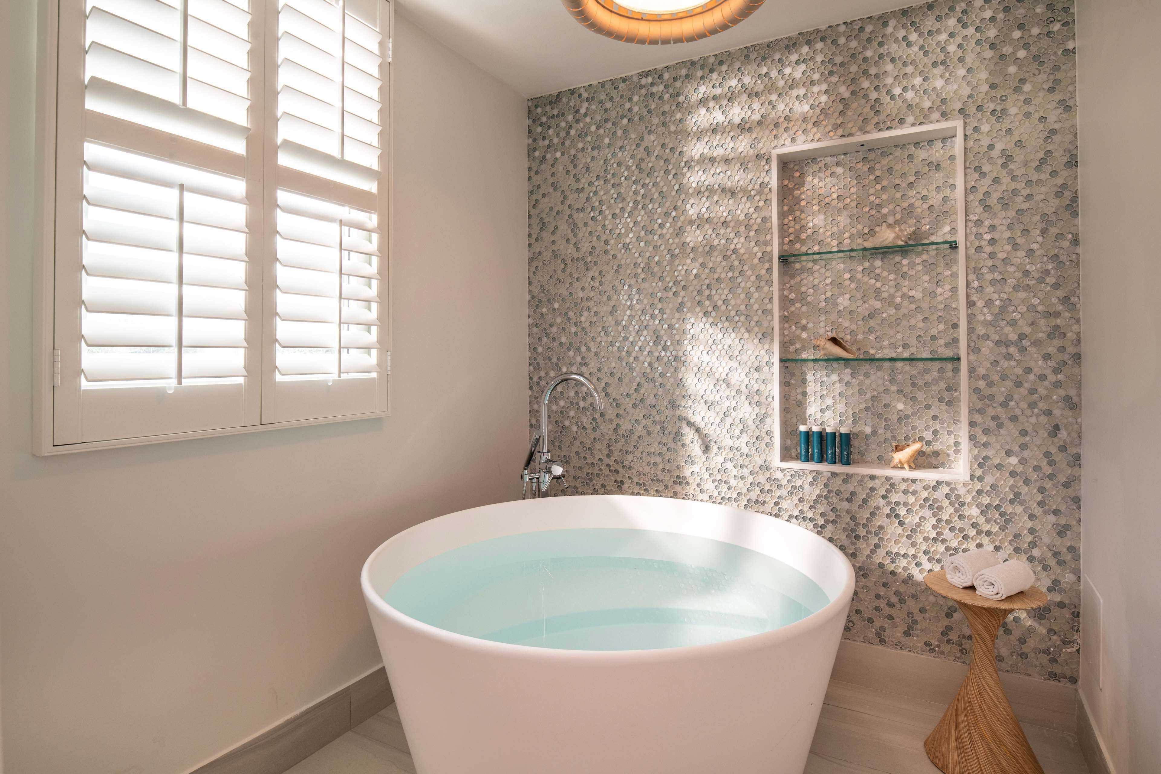 A bathroom with a freestanding bathtub, a window with blinds, a shower, and a shelf with toiletries.