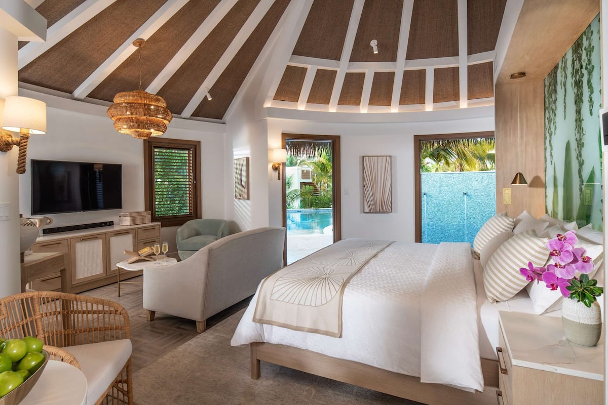 Ein geräumiges Schlafzimmer mit einem großen Bett, einem Fernseher, einer Sitzecke und Blick auf einen Swimmingpool.