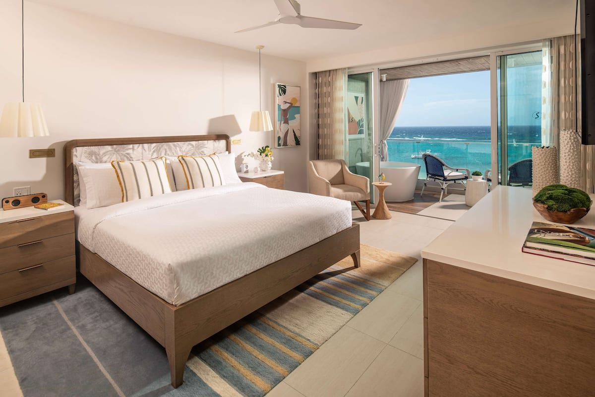 Ein modernes Hotelzimmer mit einem großen Bett, einem Schreibtisch, einem Sessel und einem Balkon mit Meerblick.