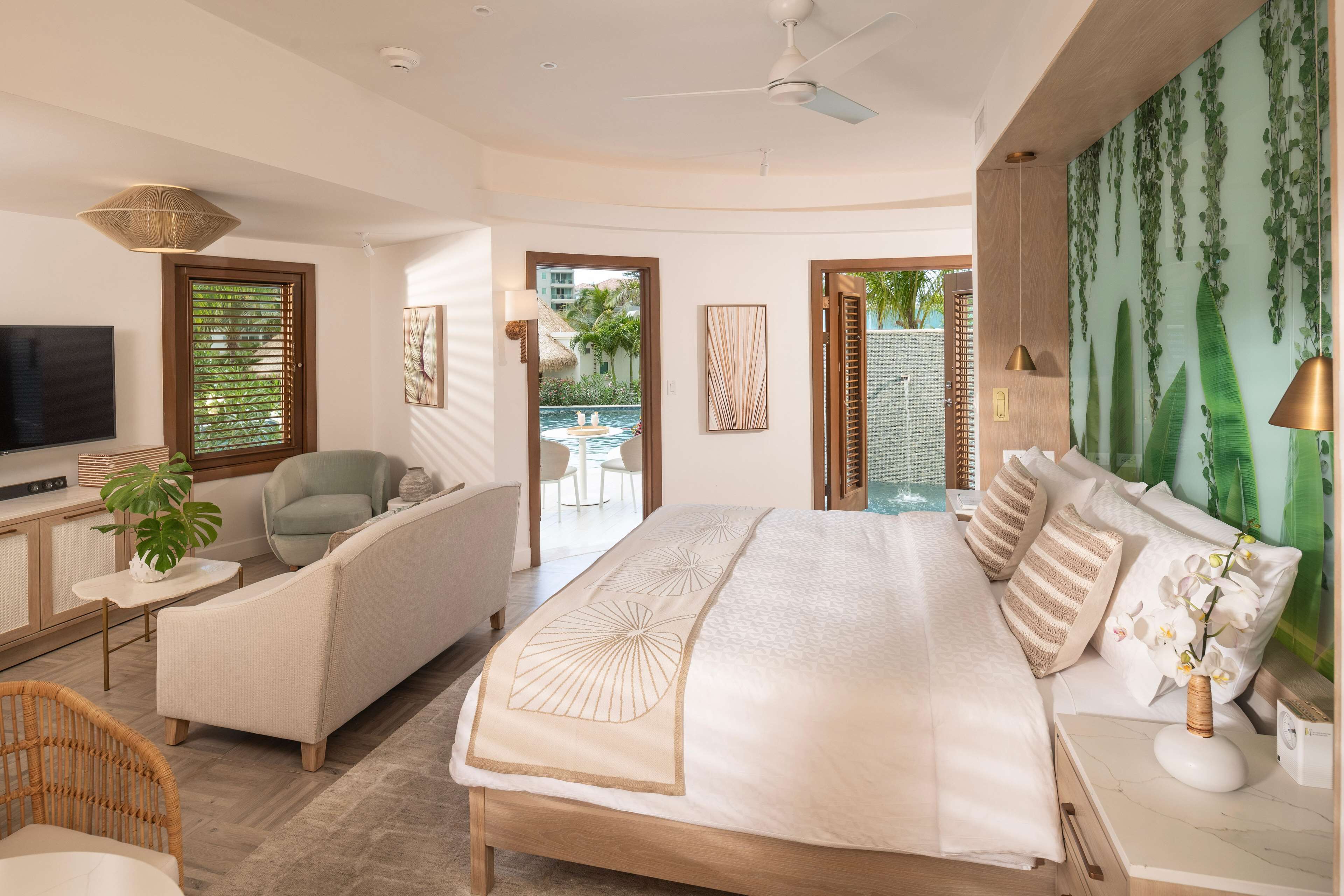 A modern hotel room with a large bed, a seating area with a sofa and armchair, a television, and a view of a pool and outdoor area.