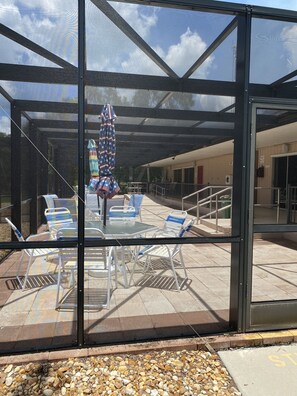 A covered outdoor seating area with tables and chairs, a patterned umbrella, and a view of the building's interior through large glass doors. - Beautiful 55+ Senior Gated Oasis: Pool, Pickleball & Small Pets Welcome! (punta gorda)