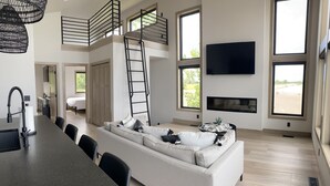 A spacious living area with a large sofa, a dining table, and a TV mounted on the wall. There is a balcony with a railing and a ladder leading to it.