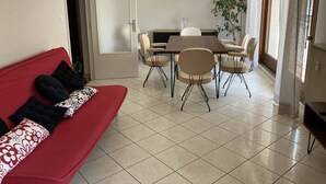 A living room with a red sofa, a dining table with chairs, and a potted plant.