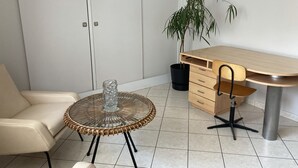 A room with a round wicker table, a beige chair, a wooden desk, a black trash bin, and a potted plant.