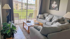 Un salon moderne avec un canapé gris, une table basse et une plante en pot. Une grande fenêtre offre une vue sur un immeuble et une pelouse.