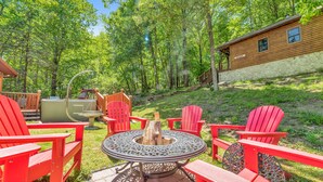 A wooden cabin with a stone foundation, surrounded by a forest. There is a fire pit with chairs and a grill.