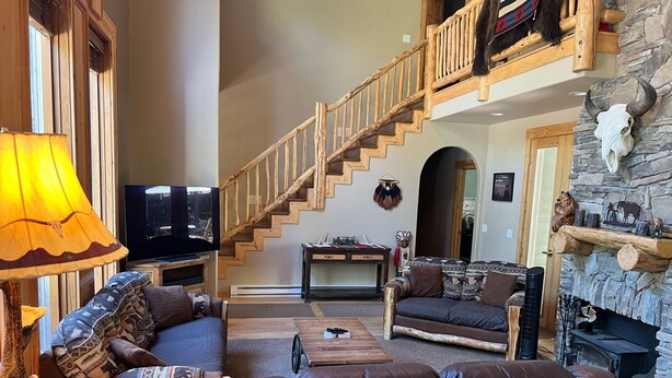A spacious living room with a wooden staircase, leather sofas, a television, and a stone fireplace. - Cozy mountain retreat with a hot tub; steps to national forest trails! (Alpine)