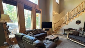 A spacious living room with a large leather sofa, a coffee table, and a TV. A wooden staircase is visible in the background. - Cozy mountain retreat with a hot tub; steps to national forest trails! (Alpine)