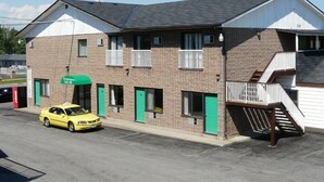 Un edificio de dos pisos con un toldo verde, un coche amarillo estacionado adelante y una escalera que lleva al piso superior.