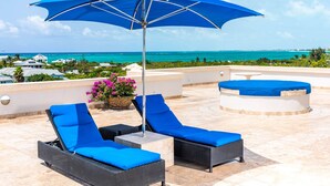 Een terras op het dak met twee lounge fauteuils en een grote blauwe parasol, met uitzicht op de kust.
