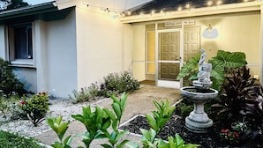 Uma casa com porta da frente, um jardim com plantas e uma fonte, e luzes de cordão.