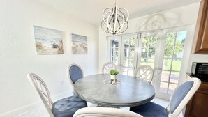 Uma área de refeições com uma mesa redonda, cadeiras brancas e um candelabro. Há duas pinturas na parede e vista para o exterior através de grandes janelas.