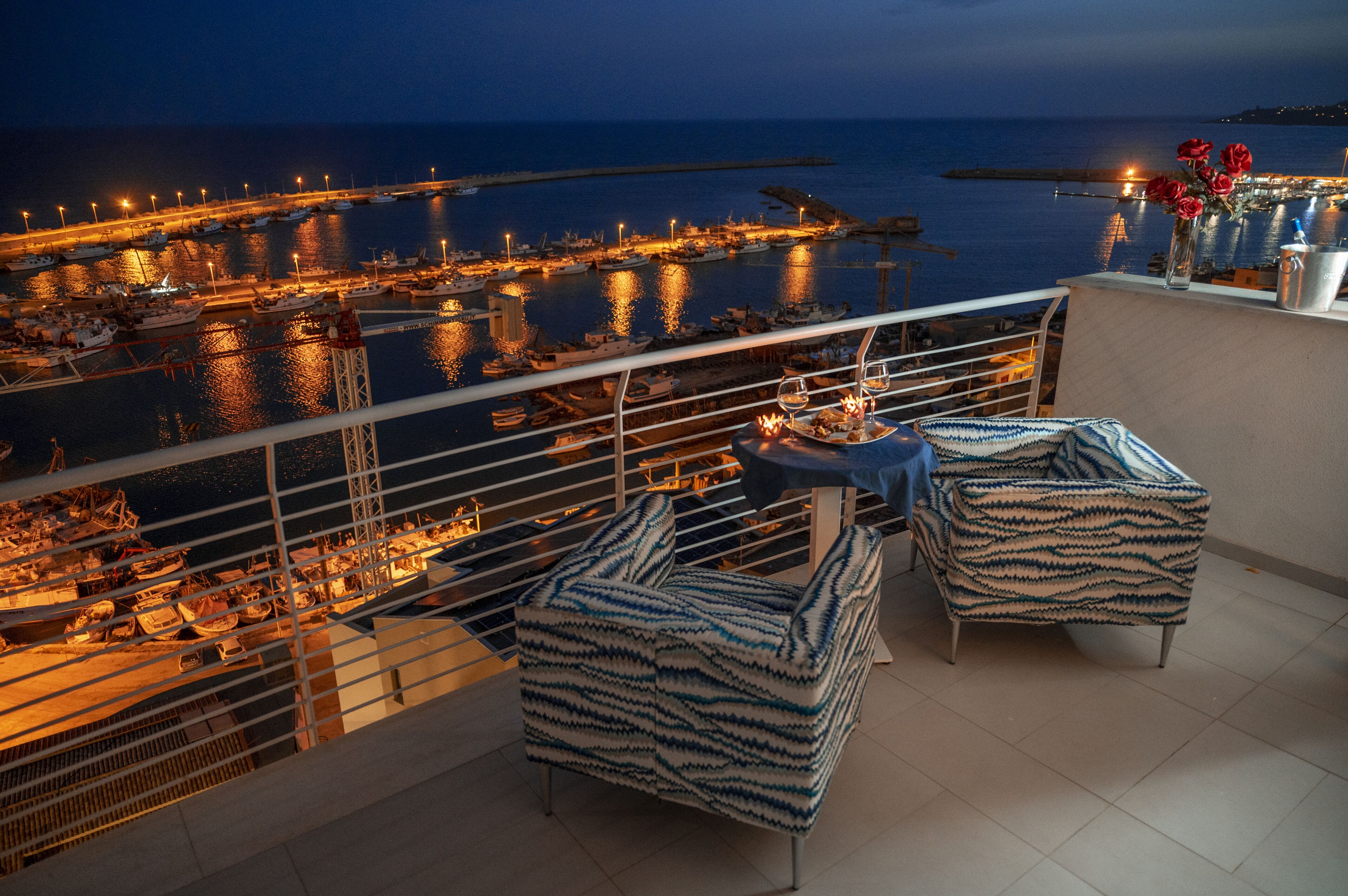 Un balcone con due poltrone decorate, un tavolino con una candela e vista su un porto turistico di notte.