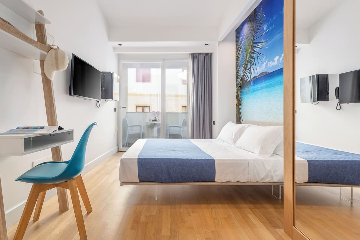 A hotel room with a bed, a desk with a chair, a TV, and a large window with a view of the sea.
