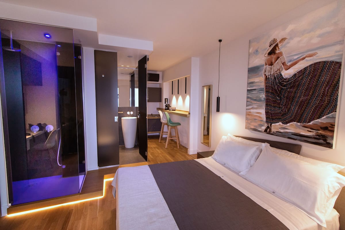 A modern hotel room with a bed, a desk, a chair, and a painting of a woman by the sea.
