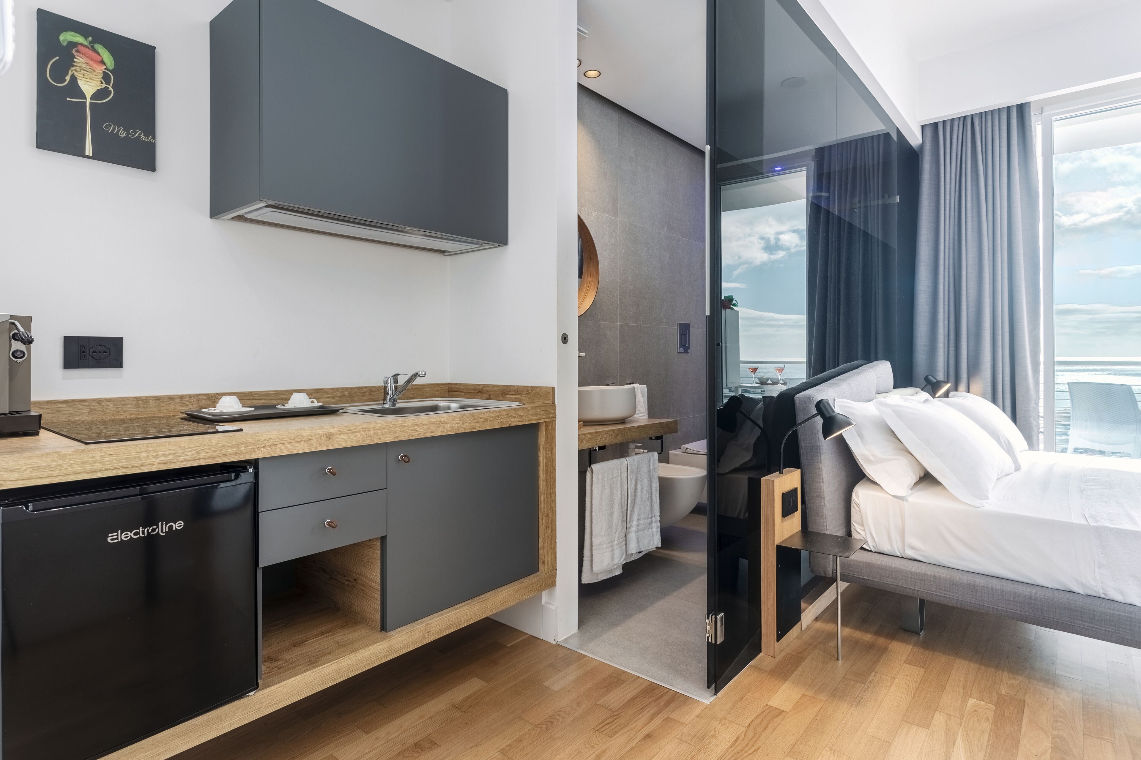 Ein modernes Hotelzimmer mit Holzboden, einem schwarzen Geschirrspüler, einem Waschbecken, einem Bett und einem großen Fenster mit Blick auf den Ozean.