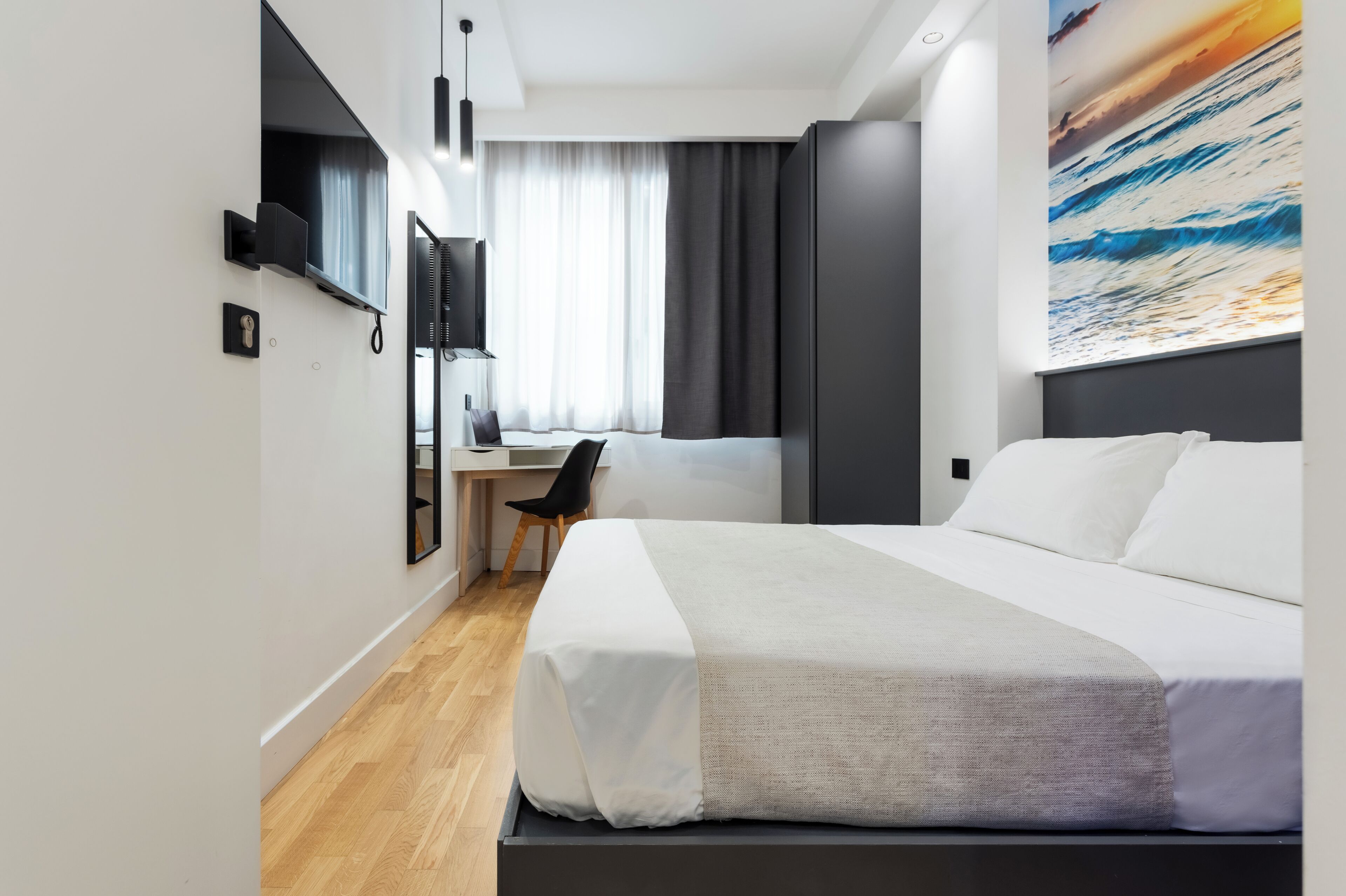 A modern hotel room with a large bed, a desk with a chair, a flat-screen TV, and a large window with curtains.