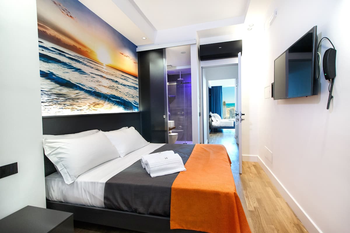 A modern hotel room with a bed, a TV, a painting, and a bathroom visible through an open door.