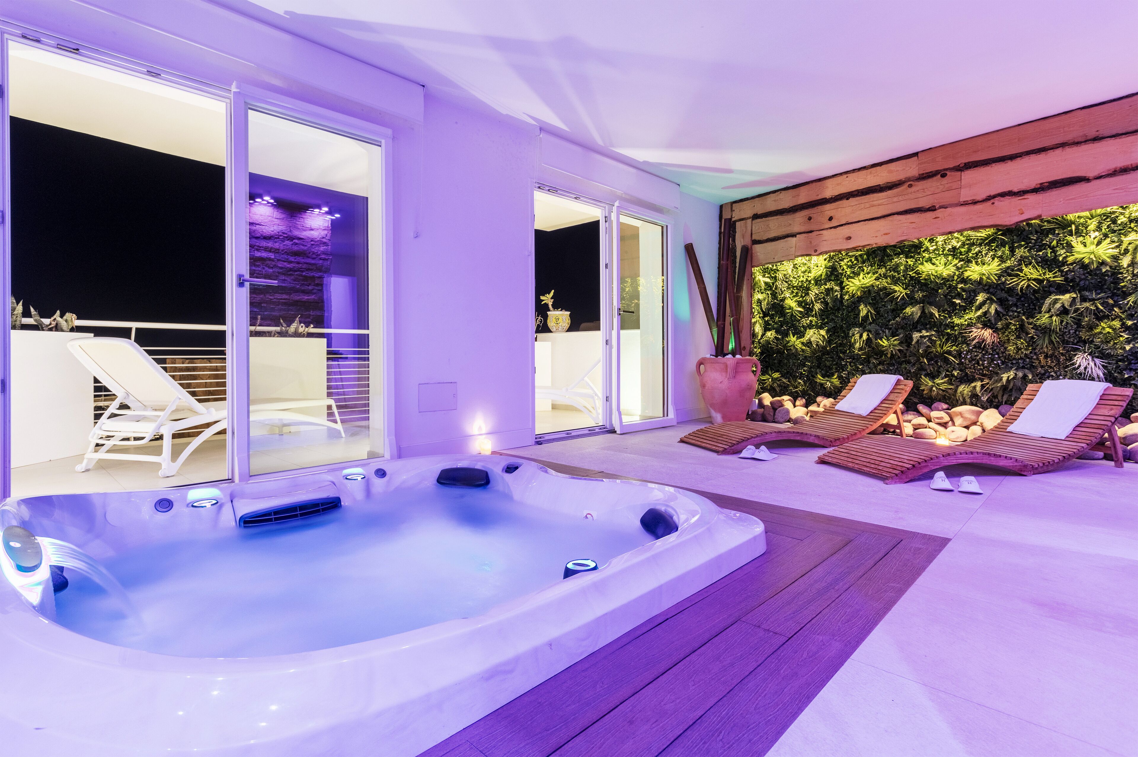 A hot tub with two lounge chairs, a wooden deck, and a glass door leading to a balcony with a view of a garden.