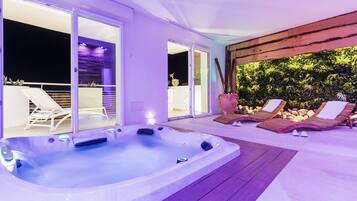 A hot tub with two lounge chairs, a wooden deck, and a glass door leading to a balcony with a view of a garden.