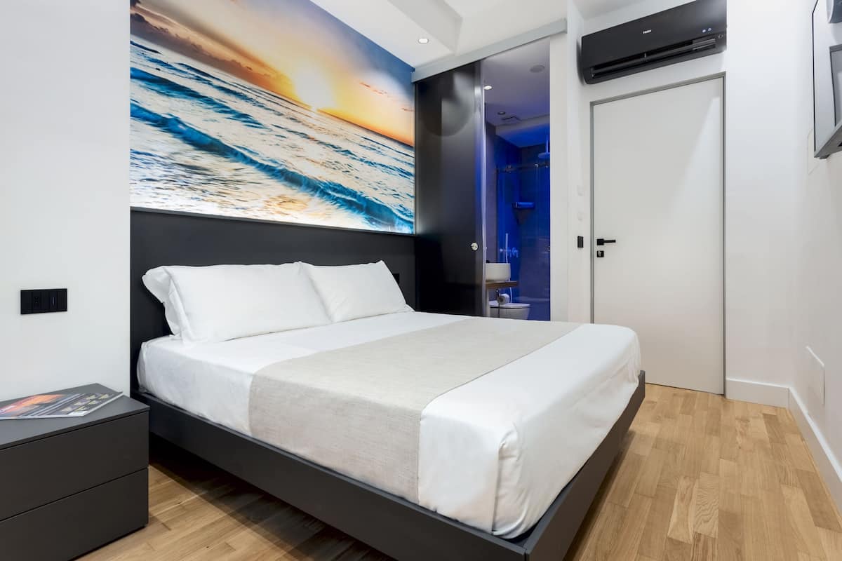 A modern bedroom with a large bed, a nightstand, a wall-mounted air conditioner, and a bathroom visible through a glass door.
