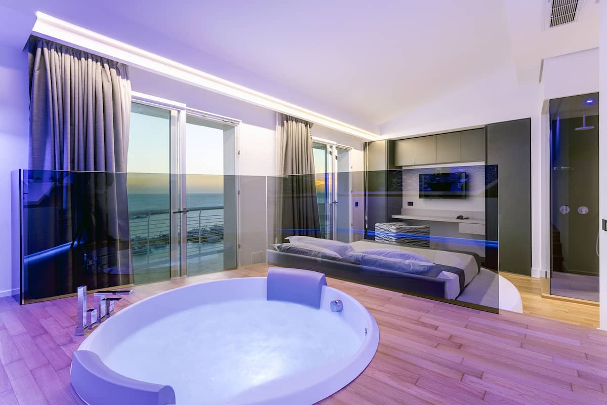 A modern bedroom with a large bed, a glass-enclosed balcony, a TV, and a hot tub.