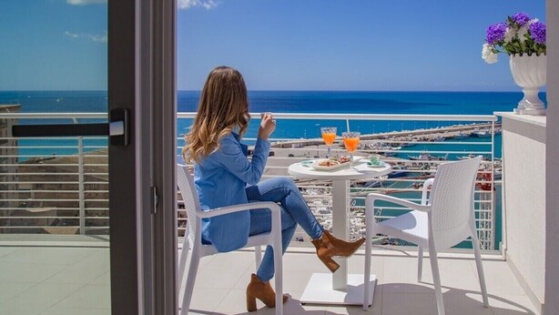 Een vrouw zit op een balkon met uitzicht op zee en een jachthaven.