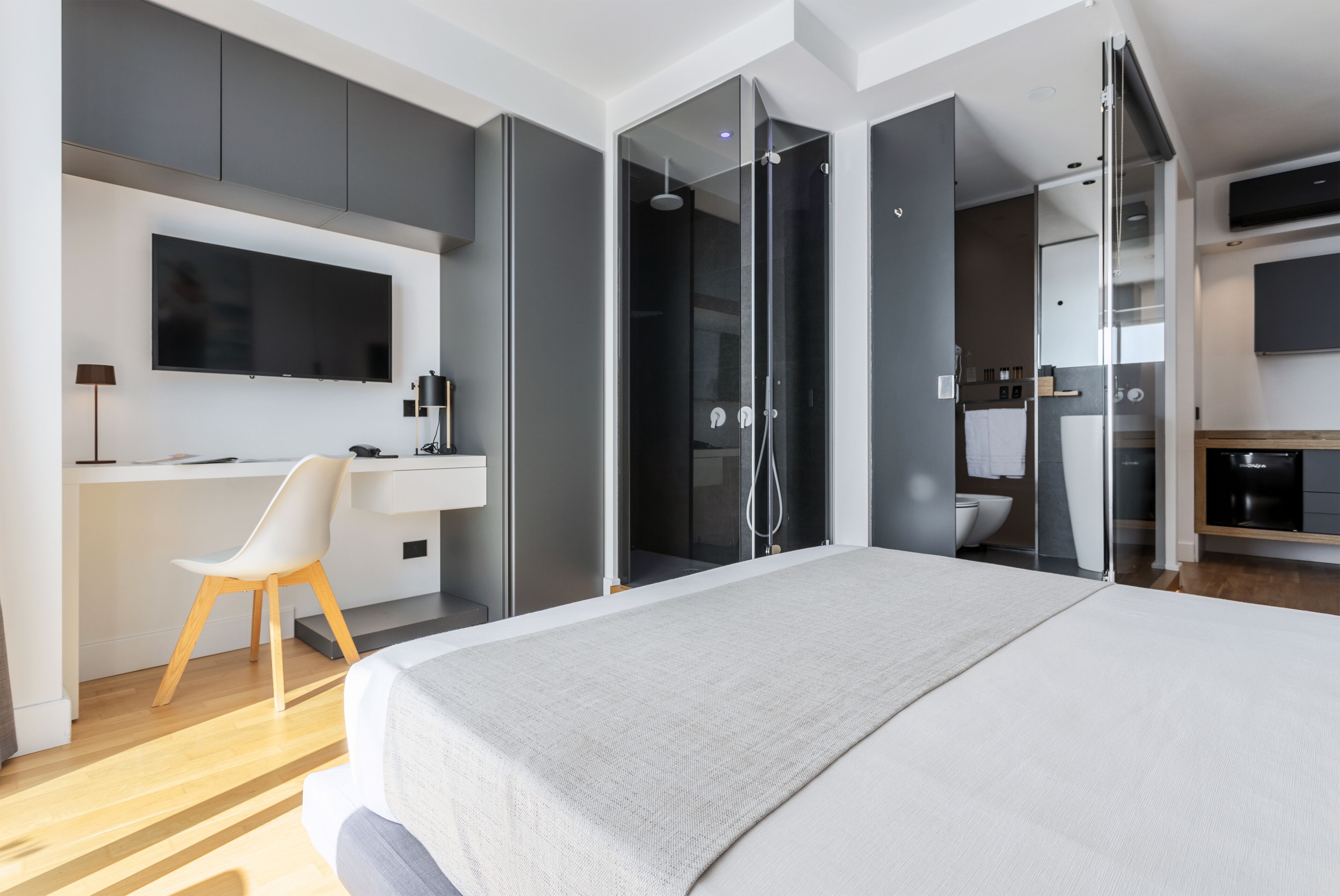 A modern bedroom with a large bed, a desk with a chair, a TV, and a walk-in shower.