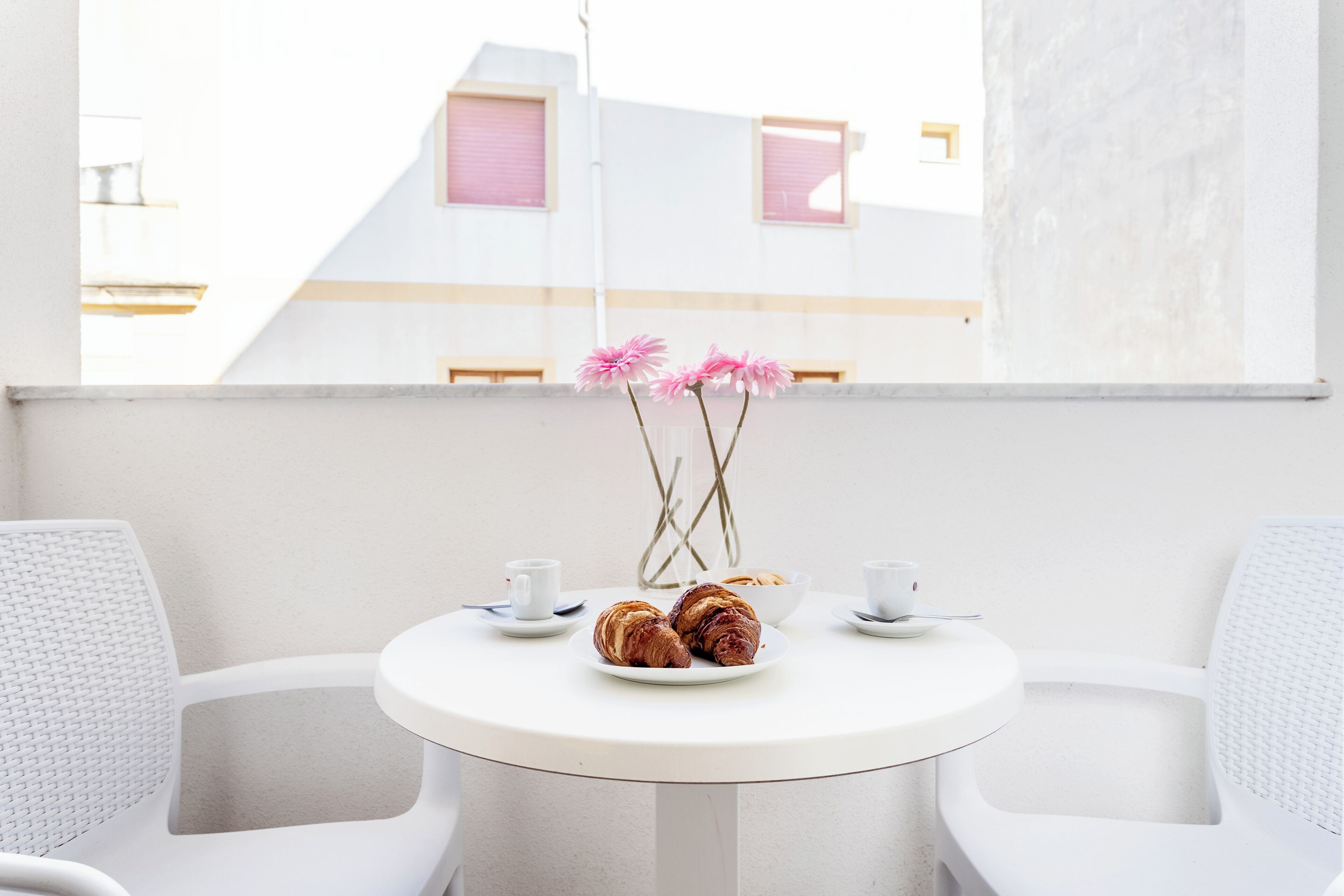 A round white table with two white chairs, a plate with croissants, a bowl, and two white cups.