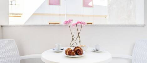 A round white table with two white chairs, a plate with croissants, a bowl, and two white cups.