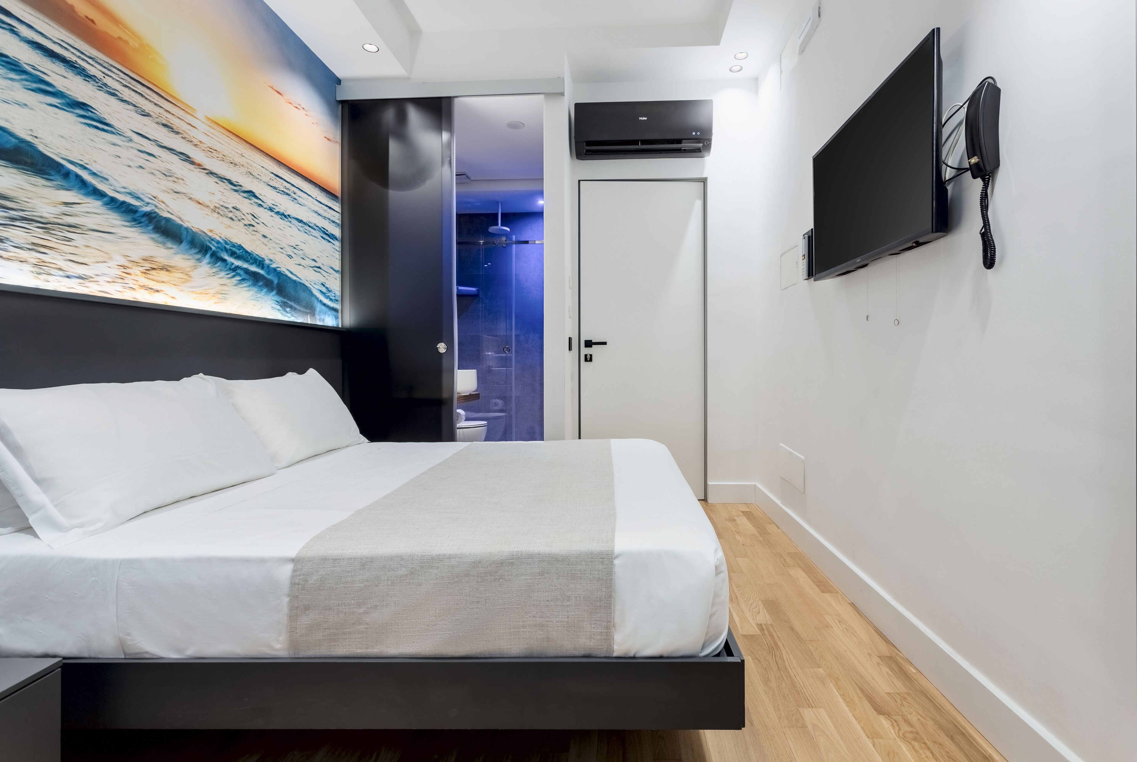 A modern hotel room with a bed, a mounted TV, and a large painting of a sunset over the ocean.