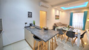 A modern kitchen with a central island, a dining area with a table and chairs, and a living space with a sofa and a window with curtains. - Tastefully furnished 3beds/3baths, 2balconies WVID1 (Gzira)