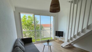 Ein modernes Wohnzimmer mit einer Couch, einem kleinen Tisch und einem Fernseher. Eine Treppe führt zu einem Balkon mit Blick auf Bäume und ein Gebäude.