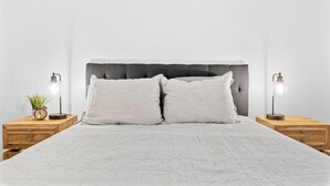 Cama bem arrumada com cabeceira estofada cinza, duas mesas de cabeceira com luminárias e um pequeno vaso com planta em uma delas.