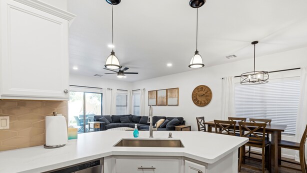 Cozinha moderna com ilha central, armários brancos e eletrodomésticos de aço inoxidável. Possui uma área de jantar com cadeiras de madeira e uma mesa, além de um ventilador de teto.