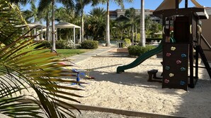 Um parquinho com escorregador e área de escalada, cercado por palmeiras e uma piscina.