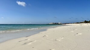 Uma praia arenosa com pegadas que levam à água, céu claro e edifícios ao longe.