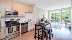 A modern kitchen with stainless steel appliances, wooden cabinets, and a dining area with a large window. - Chic 1 bed Apt + Rooftop & Gym - Downtown Stamford (Stamford)