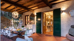 A dining area with a round table set for four, surrounded by white chairs. A chandelier hangs above, and a stone wall with greenery is visible in the background. - Villa Arsina in Lucca (Lucca)