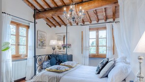 A bedroom with a wooden ceiling, a chandelier, a bed with white bedding, a nightstand, and a window with curtains. - Villa Arsina in Lucca (Lucca)
