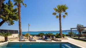 A pool area with lounge chairs, palm trees, and a view of the sea. - Beachfront Villa Tota with Pool (Kaštel Štafilić)