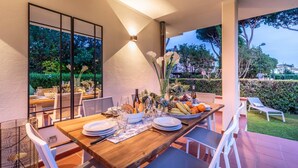 A dining area with a wooden table set for a meal, surrounded by white chairs, with a view of a garden and a hedge in the background. - Regina Beach Villa by the Beach in Marina di Piet (Camaiore)