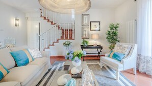 A spacious living room with a staircase, a white sofa with blue cushions, a white armchair, a large woven ceiling light, and a small table with decorative items. - Regina Beach Villa by the Beach in Marina di Piet (Camaiore)
