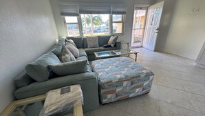 A living room with a sectional sofa, a glass coffee table, and a patterned ottoman.