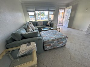 A living room with a sectional sofa, a glass coffee table, and a patterned ottoman.