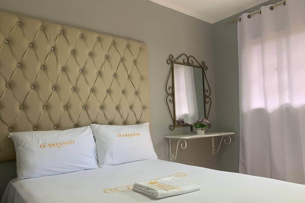 A hotel room with a tufted headboard, two white pillows with 'GLAMOUR HOTEL' embroidered, a bed with a white bedspread, a folded towel, a bedside table with a mirror, and a white curtain.