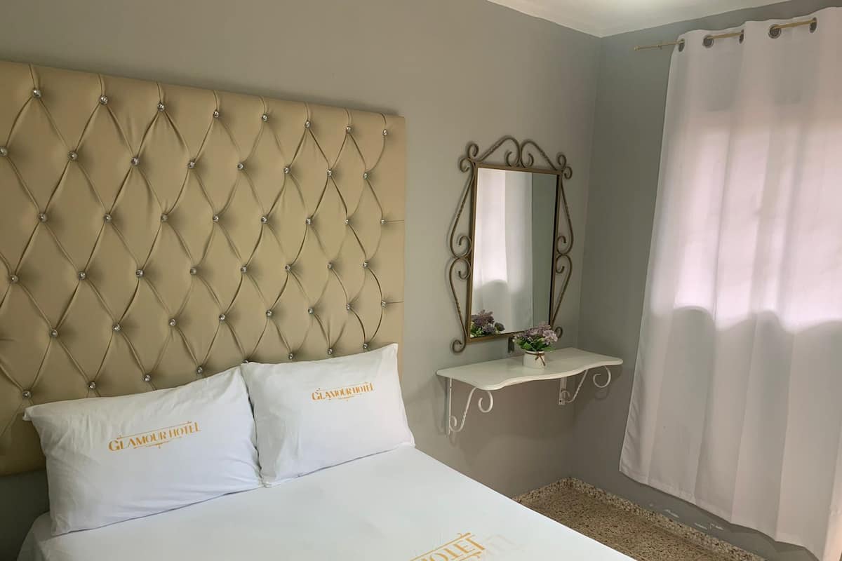 A hotel room with a tufted headboard, a bedside table with a mirror, and a white bedspread with the hotel name.