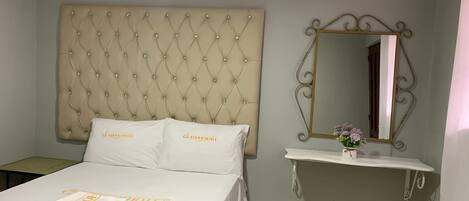 Una habitación de hotel con un cabecero capitonado, una cama con ropa blanca, una mesita de noche con un jarrón de flores y un espejo decorativo.